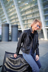 Fototapeta premium Trendy Woman in Leather Jacket Sitting Outdoors by Modern Architecture..