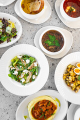 A variety of dishes displayed on a gray textured table. Plates include salads, soups, and a hearty stew, showcasing colorful vegetables and garnishes.