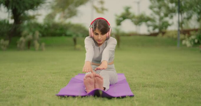 Indian young adult gen z woman lady sitting on yoga mat do stretch warm up toe touch asana in fresh air outdoor garden cute teen age female girl learn good self health care habit day time at park