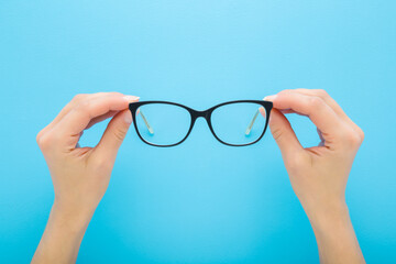 Young adult woman hands holding eyeglasses with dark black plastic frame on light blue table...