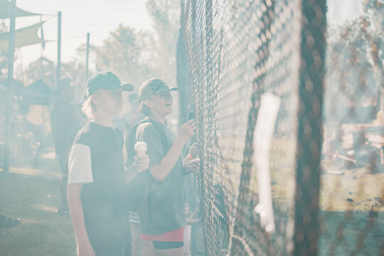 Teenagers watching vehicle burnout competition through chain mesh fence with smoke haze from burning