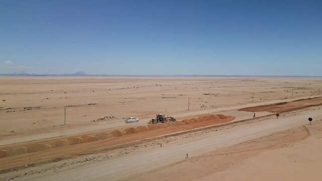 Tractor grader levels a road in the vast Namib Desert, Namibia, with endless sand and clear blue skies, aerial medium orbit