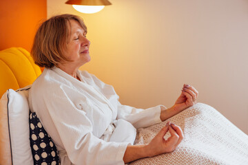 An elderly woman in a white coat is sitting in bed and meditating
