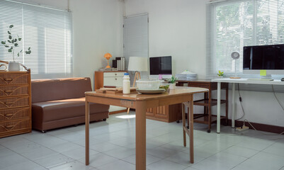 Wooden table with breakfast leftovers in the foreground, brown leather couch, computer desk with desktop computer, white venetian blinds filtering sunlight in a messy home office