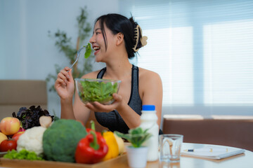 Smiling asian sportswoman enjoying a healthy fresh salad at home, surrounded by a variety of vegetables and fruits, promoting a healthy and active lifestyle