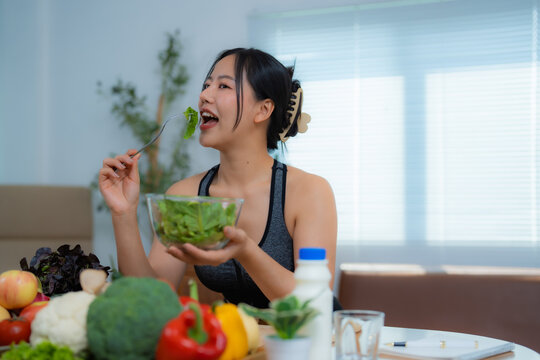 Happy asian fitness girl eating fresh green salad after workout at home, enjoying healthy food, promoting healthy lifestyle for weight loss