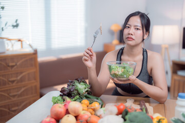 Young woman wearing sportswear holding a bowl of salad with an unhappy expression, sitting at a...