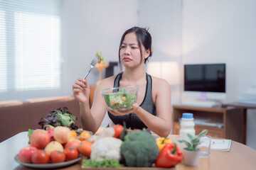 Young woman with an unhappy expression eating a salad at home, surrounded by fresh vegetables and fruits, while following a strict diet plan for weight loss and a healthy lifestyle