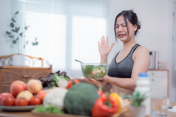 Young woman wearing sportswear is refusing to eat healthy salad while crying and gesturing with her hand in a domestic kitchen with many vegetables and fruits on the table