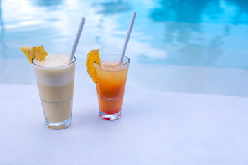 Close-up of two colorful delicious cocktails on the side of the pool. Glasses with cool drinks and pieces of fruit near the clear blue water.