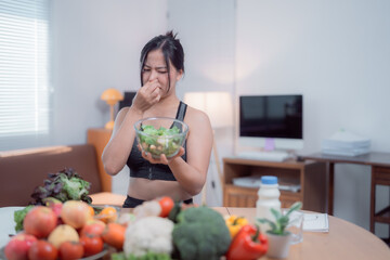 Young sportswoman pinching her nose in disgust while holding a bowl of salad, surrounded by fresh produce on a table in her apartment