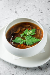 A bowl of rich, steaming soup with mushrooms and vegetables, garnished with fresh parsley, served on a minimalist plate against a textured gray background.