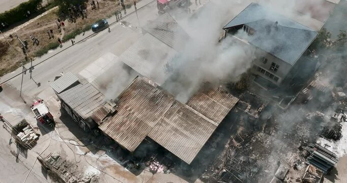 Aerial footage  burning factory as fire trucks and crews respond. Dense black smoke pours from structure, highlighting an urgent emergency situation.Building fire incident in summer