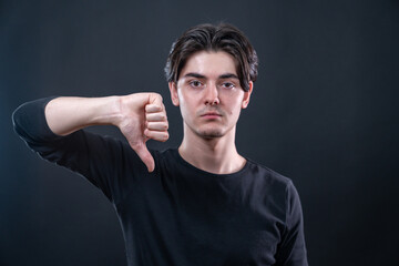 Young man showing a thumb down sign with a serious expression on isolated black background. The focus is on the hand sign.