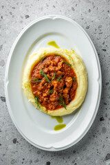 A delicious serving of creamy mashed potatoes topped with savory meat sauce, garnished with fresh herbs, on a sleek white plate. The background features a textured, light surface.