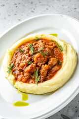 A delicious serving of creamy mashed potatoes topped with savory meat sauce, garnished with fresh herbs, on a sleek white plate. The background features a textured, light surface.