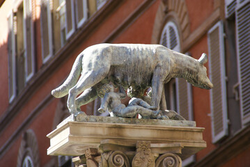The iconic Capitoline Wolf statue, depicting Romulus and Remus suckling, stands against a warm, sunlit building. A symbol of Rome's founding, captured in a classic shot.