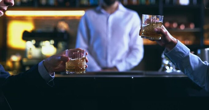 Close up of whisky glasses hitting together at a bar counter. Friends cheering at a bar counter. Close up of two men hands clinking drink glasses in luxury restaurant bar. 
