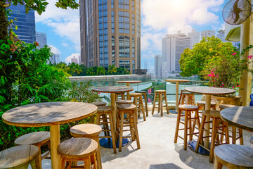 Vibrant outdoor view seating area features rustic wooden tables and stools set against a backdrop of modern skyscrapers under a clear blue sky.