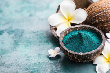 A vibrant blue-green powder in a coconut bowl, surrounded by tropical flowers, set against a textured teal background.