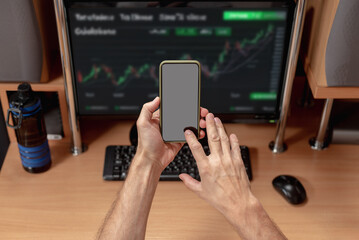 Male stock exchange trader using smartphone analyzing stock market investment. Nervous trader at home, man watching charts on monitor