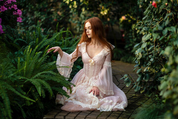 Beautiful red head girl in pink  vintage dress  standing near green plants and fern. Art work of...