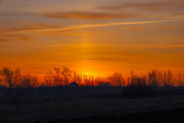 This photograph captures a breathtaking sunset over a rural landscape. The sun sets behind the horizon, flooding the sky with vibrant shades of orange and yellow.
