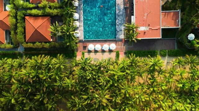 Overhead tropical resort scene showing expansive swimming pool, white sunshades, palm trees, orange roofed bungalows nestled among verdant greenery