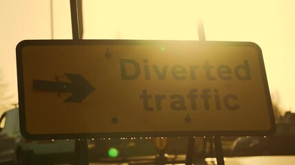 Diverted traffic sign standing on illuminated roadway. Highway sign shadow in early sun. Road detour board appearing as dark outline in golden morning haze. Traffic redirection panel blending into