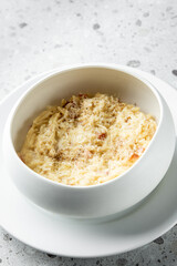 A creamy risotto served in a modern white bowl, garnished with grated cheese and a sprinkle of pepper, on a textured gray surface.
