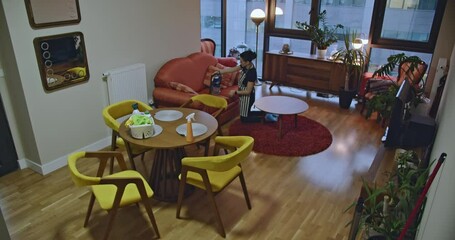 A woman vacuums a sofa in a small apartment. Cleaning supplies are visible on a nearby table.
