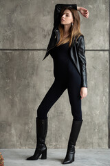 Fototapeta premium Confident woman in a black bodysuit and leather jacket, posing against a gray concrete wall, wearing black boots