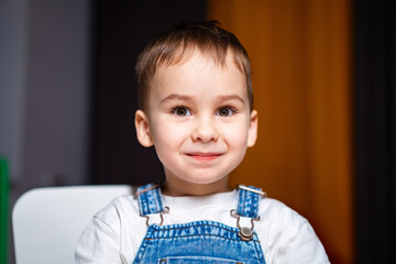 Smiling child in denim overalls at home. A young boy with short hair and bright eyes smiles while...