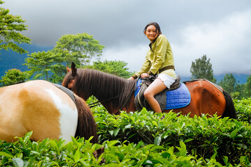 Happy smiling Southeast Asian teenage girl riding a horse on a rural mountainside tea farm in the morning during vacation for refreshing