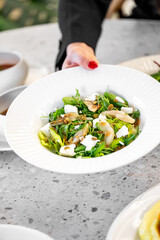 A fresh salad featuring greens, feta cheese, and herbs is being served on a white plate. Hands gently present the dish, showcasing its vibrant colors and textures.