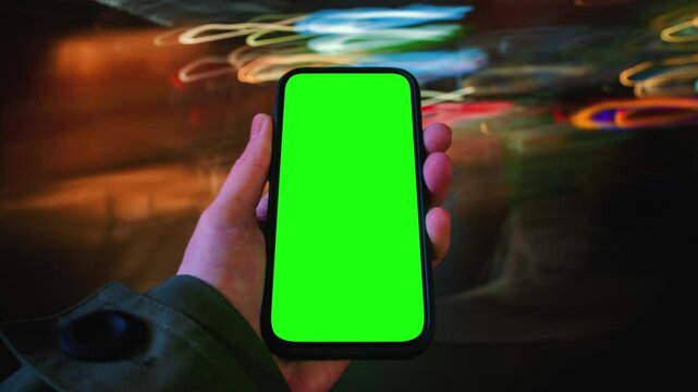 Close-up time-lapse hyperlapse of male hand holding modern smartphone keylight green screen gadget walking city street at night, fast motion blurry light trails glow, pov phone screen with chroma key