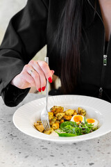 A close-up of a woman’s hand holding a fork, about to eat a plate of colorful food featuring roasted potatoes, salad, and eggs. The stylish setting enhances the appetizing presentation.