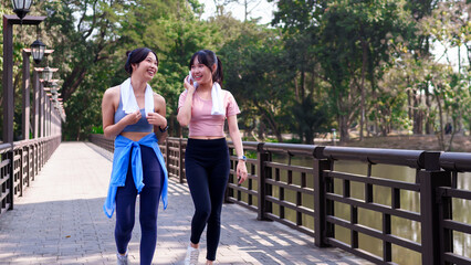 Two young Asian women walking and chatting on a wooden bridge after exercising together in a scenic park