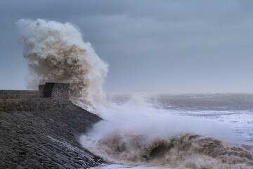 A huge winter storm hitting the coast of the UK. Giant waves crash into a breakwater, with a lighthouse. The sky is grey and moody