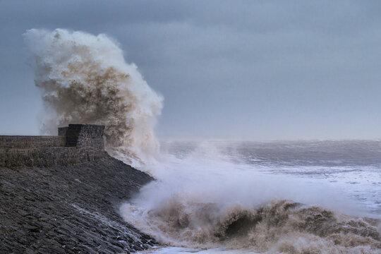 A huge winter storm hitting the coast of the UK. Giant waves crash into a breakwater, with a lighthouse. The sky is grey and moody