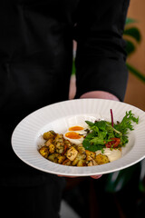 A chef presenting a plate of beautifully arranged roasted potatoes garnished with herbs, accompanied by a fresh salad and a creamy sauce, set against a blurred background.