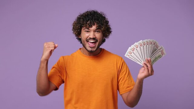 Happy young Indian man wears orange t-shirt casual clothes hold fan of cash money in dollar banknotes do winner gesture celebrate isolated on plain pastel light purple background. Lifestyle concept