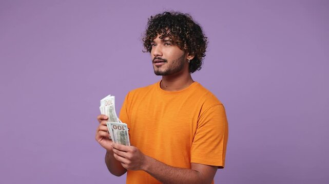 Happy accountant young Indian man he wear orange t-shirt casual clothes hold in hand count stack of cash money in dollar banknotes isolated on plain pastel light purple background. Lifestyle concept