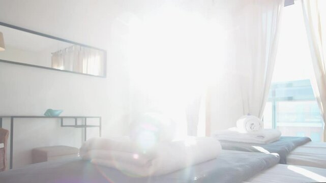 Freshly folded white towels rest on soft gray blankets in a sunlit modern bedroom with large windows and sheer curtains, creating a tranquil and cozy atmosphere