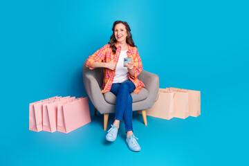 Happy mature woman in a stylish outfit sitting with shopping bags on a blue background while using...