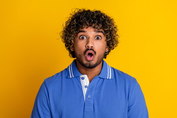 Surprised young man with curly hair wearing a stylish blue polo shirt against a vibrant yellow background