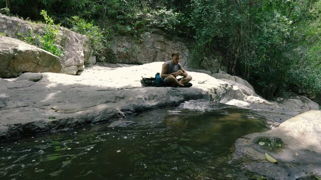 A tourist sits next to a mountain stream at a rest stop and eats