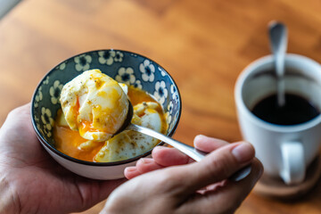 A close up of a hand holding a spoonful of soft boiled egg with runny yolk with a coffee for breakfast
