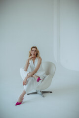 Fashion portrait.  Woman in a white suit and pink shoes sits on a white chair on a white background. young blonde woman. mood of business style, fashion, design and minimalism. There is space for text