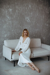 A beautiful young girl in a white elegant outfit is sitting on the sofa. Her look conveys the mood of luxury, wealth, style and fashion. Fashion portrait of a blonde girl. Old money